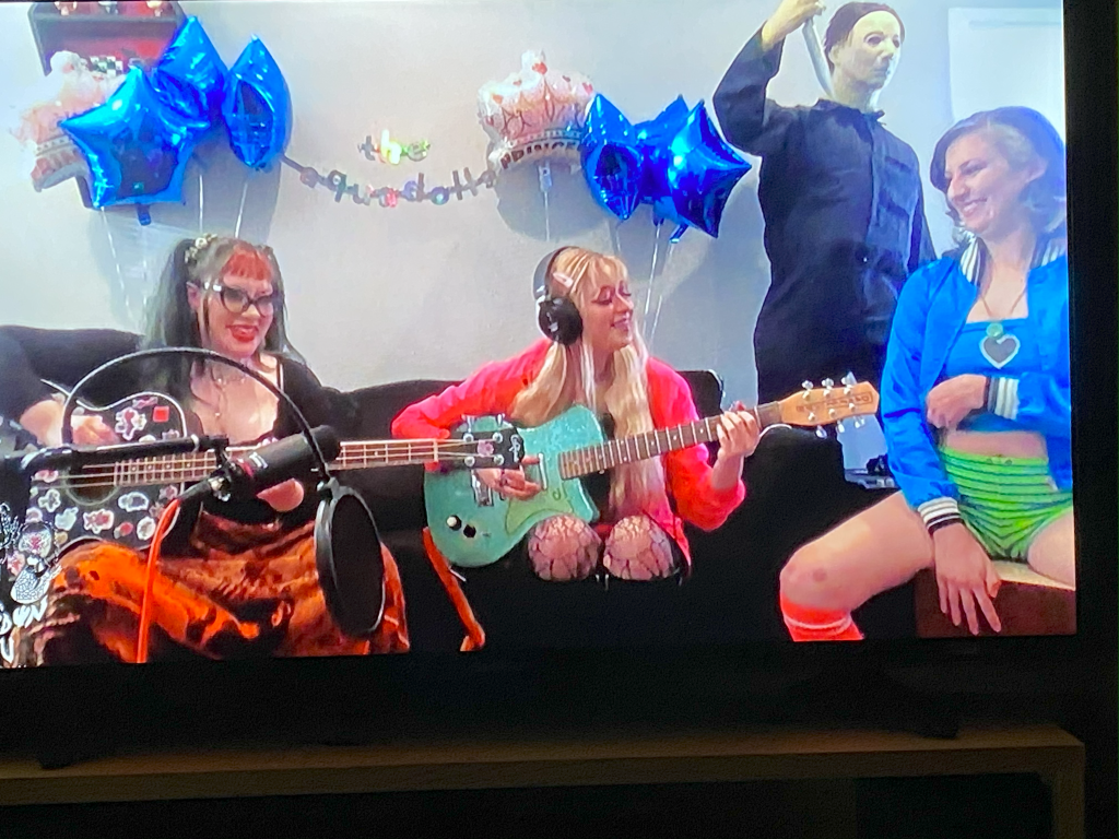 The Aquadolls, Keilah, Melissa and Jackie sit on a couch, smiling and performing.