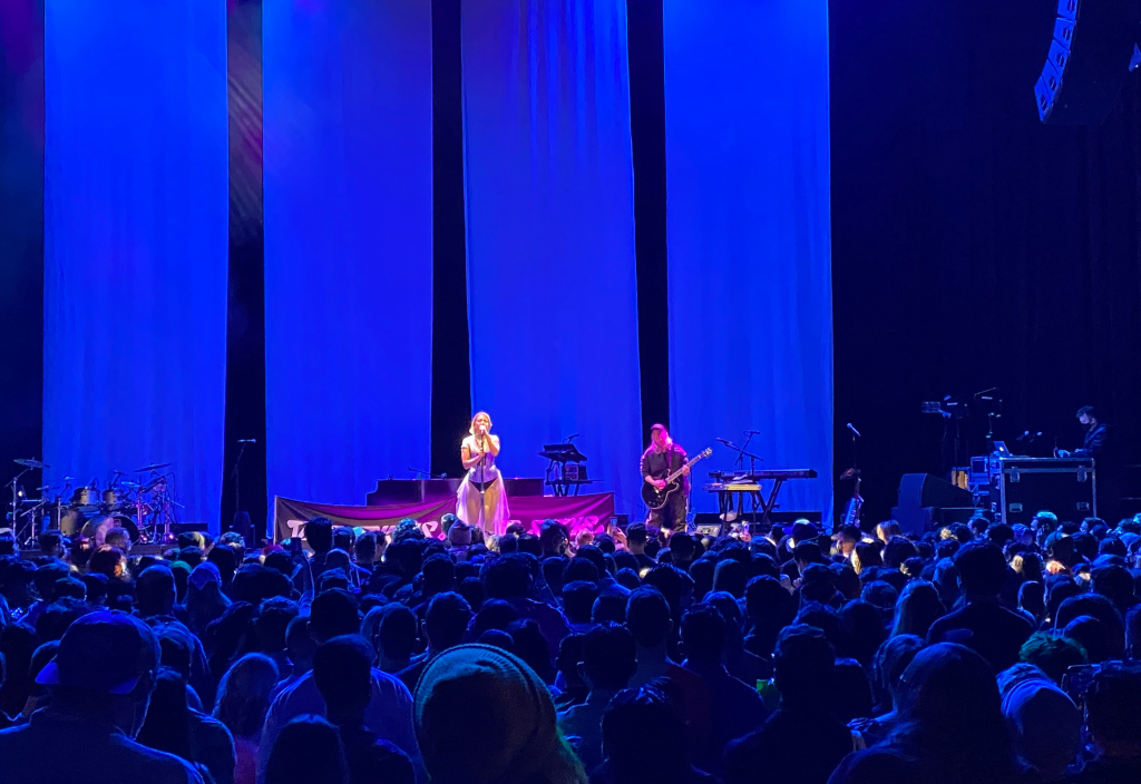 Tove Styrker performs at the Masonic auditorium. She's wearing a purple corset, with a flowing white tulle skirt partially covering her bare legs. She has one accompanist who is playing the guitar.