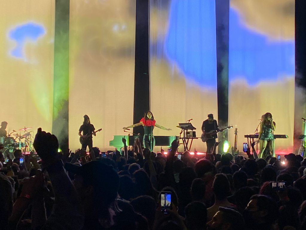 Maria Diamandis and band on stage. Marina stands with arms spread and the other members of the band play and sing on either side.