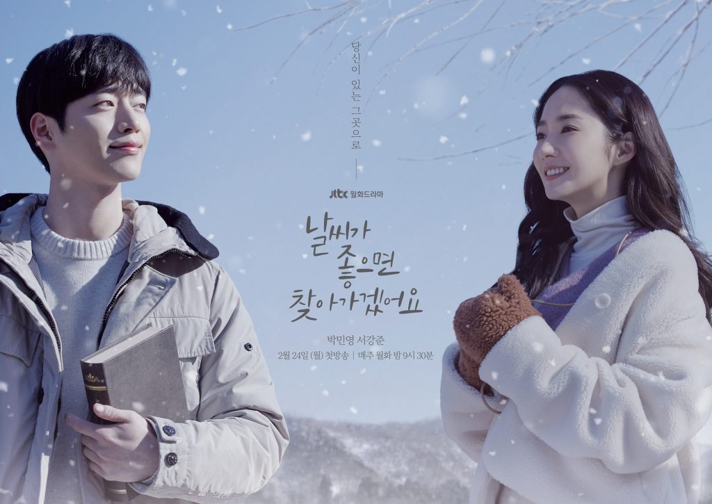 Still from the drama When the Weather is Fine. It features a 3/4 view of the two main characters dressed in light colored winter jackets - an attractive man and a woman who appear to be around 30 years old. Behind them is a pale blue sky with snowflakes. A little bit of snow covered forest is visible behind them, and some bare tree branches hang down from above. Male lead Eun-seop is on the left, his body towards the viewer, but his head is turned to his left in the direction of female lead Hae-won. Eun-seop is wearing a half smile, and holds a hard-cover book in his arm. Hae-won is smiling, and stands at an angle in the direction of Eun-seop. She has a fuzzy white coat, and brown furry mittens.